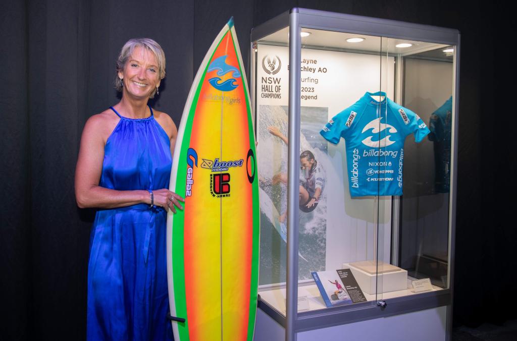 Surfing inductee Layne Beachley next to surfboard, and display case containing photo of Layne surfing and mounted swim shirt.