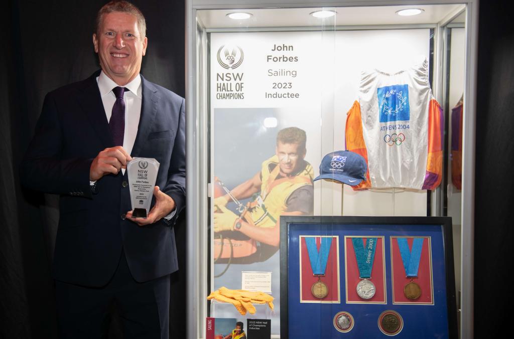 Sailing inductee John Forbes holding award, next to display case containing photo of John on sailing rigging, and Olympic games uniforms and medals.
