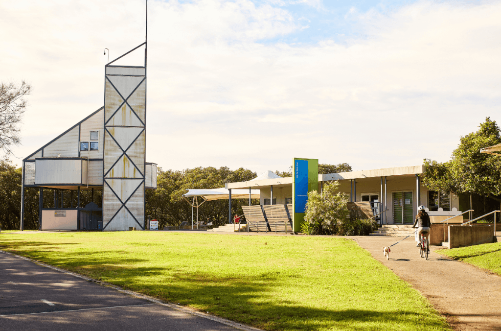 A cyclist walks a dog along a paved path beside a modern park building and tall tower, with grass lawns, trees, and shade sail structures nearby.