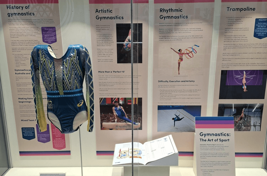 A museum display case features a blue and gold gymnastics leotard alongside informational panels about artistic, rhythmic, and trampoline gymnastics, with photos of athletes and an open booklet below.