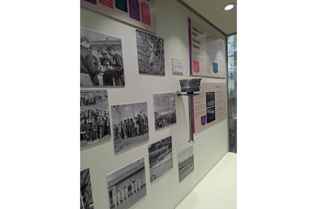 A museum wall display presents black-and-white photographs and text about the 1956 Melbourne Olympic Games, alongside a ceremonial torch mounted in a glass case.
