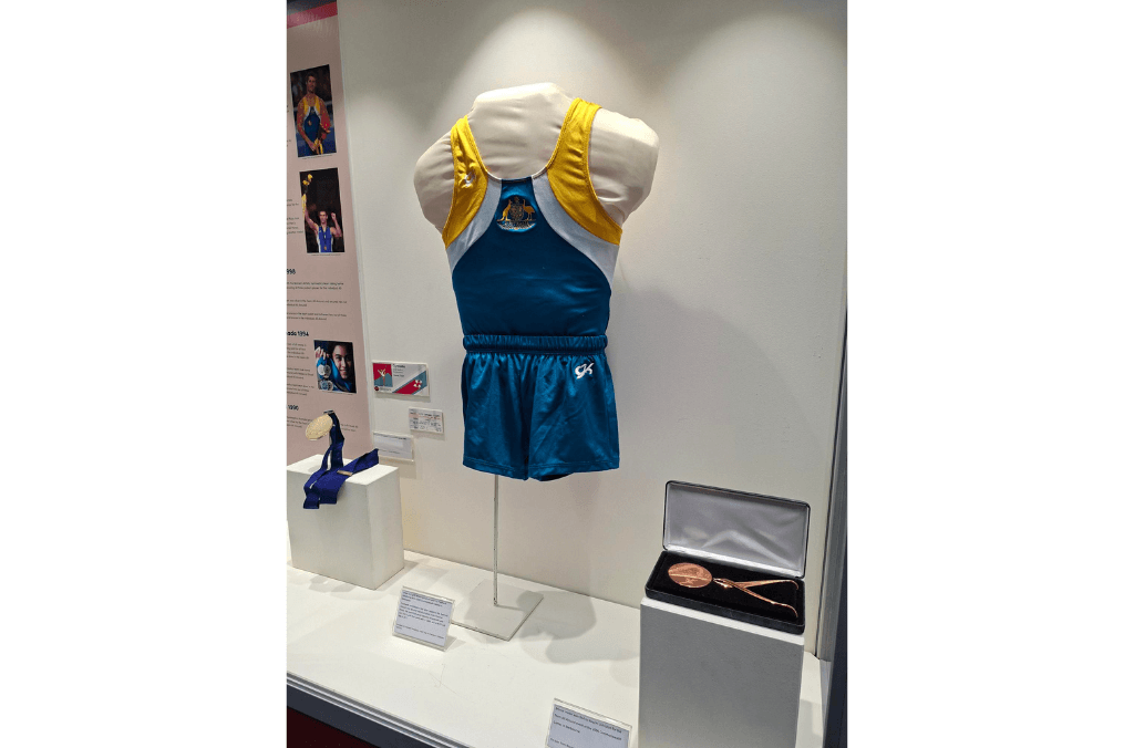 A museum display showcases a blue and yellow gymnastics uniform on a mannequin alongside medals, a commemorative spoon in a case, and informational panels with athlete photos.