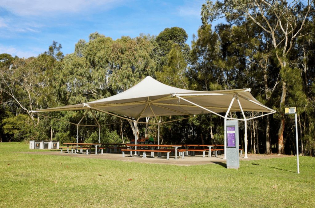 A covered picnic area with long tables sits on a grassy clearing, surrounded by tall trees under a blue sky.