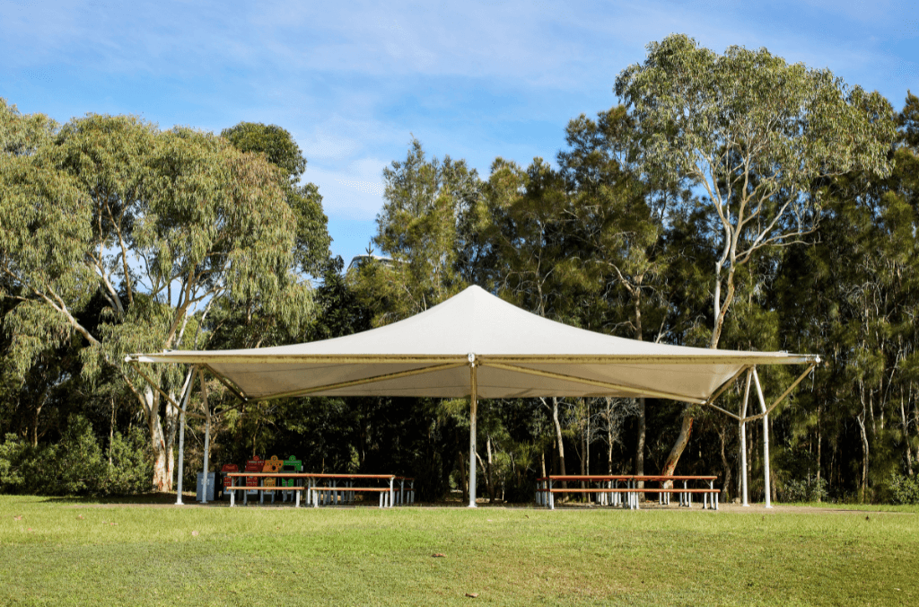 A large shade sail shelters picnic tables in a grassy park clearing, with tall trees forming a dense green backdrop under a bright blue sky.
