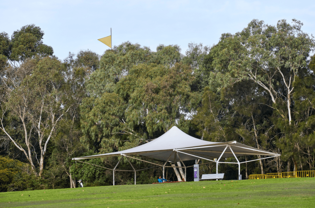 A shade sail structure shelters picnic tables on a grassy slope surrounded by trees, with a flag visible above the treeline in the background.